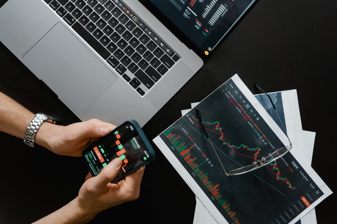 about-01 Hands using smartphone beside laptop with stock charts, showcasing digital trading.